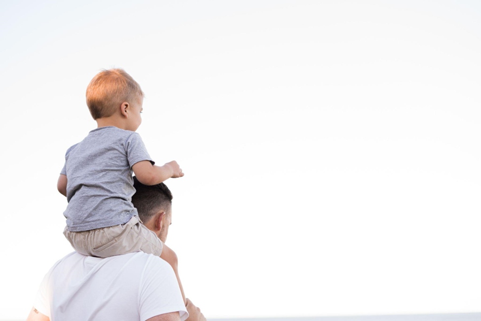 Alt Text: A man stands with his infant son on his shoulders looking at the ocean